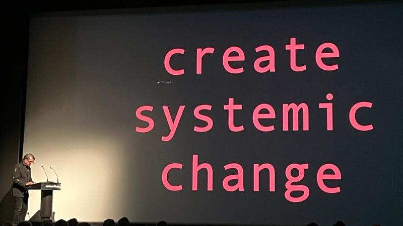 Nusrat Durrani delivers keynote in front of screen that reads 'create systemic change'