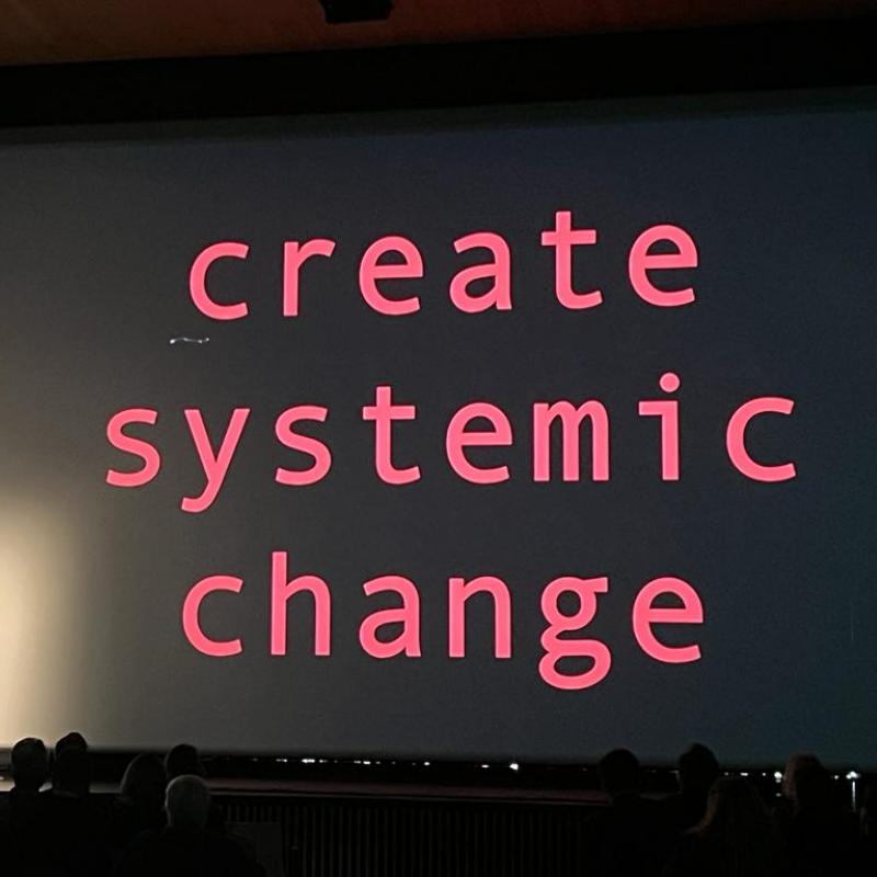 Nusrat Durrani delivers keynote in front of screen that reads 'create systemic change'