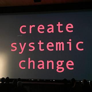 Nusrat Durrani delivers keynote in front of screen that reads 'create systemic change'