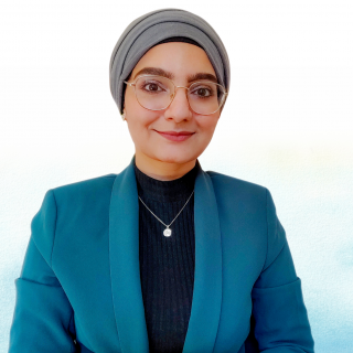 Zainab Kidwai smiles. She wears glasses, a blue blazer, and a grey headscarf.